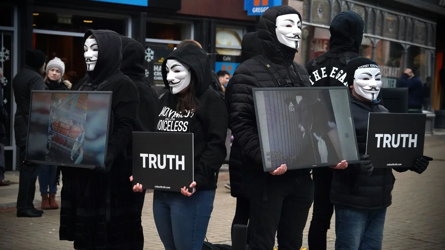 Aktivist:innen von Anonymous for the Voiceless stehen mit Masken und Bildschirmen in einer Cube-Formation.